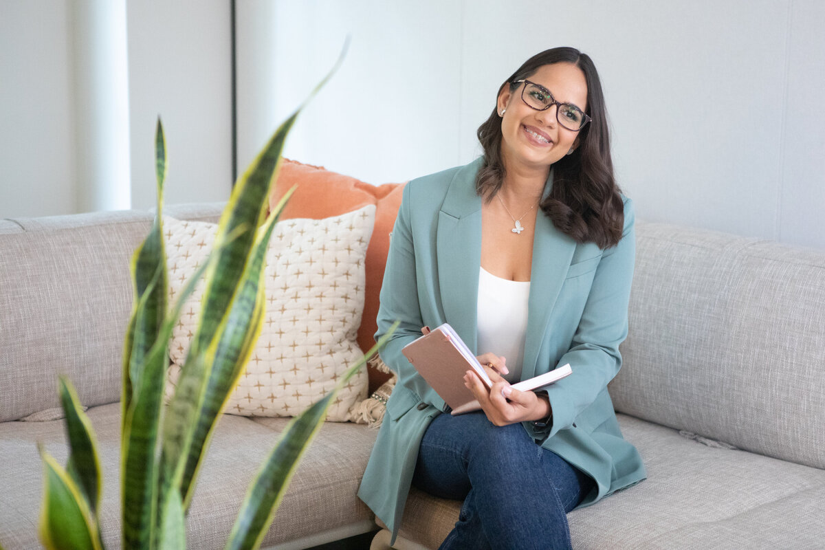Latina young woman in a sea foam green blazer smiles and holds a tan notebook on a grey couch with orange and cream throw pillows New York branding photographer Milestone Images