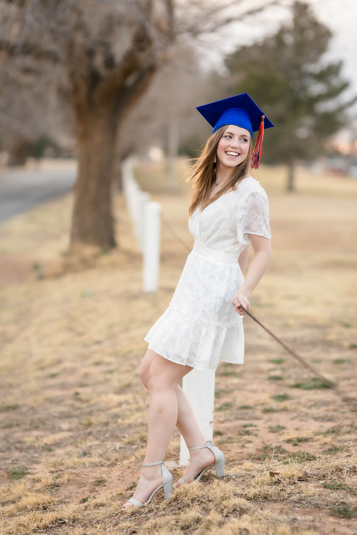 Woman college mini session with woman wearing a white dress  and shoes and wearing a blue SPC graduation cap with tassel.  She is leaning on a wire fence bordering Levelland Country Club and smiling off camera