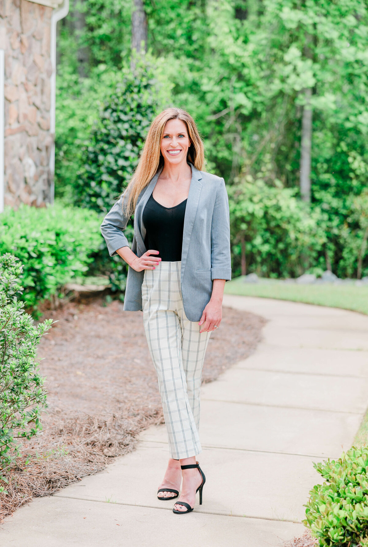 A woman stands on a sidewalk, smiling at the camera. She is wearing a gray blazer, black top, and white plaid pants paired with black heels. Greenery and a stone wall are visible in the background.