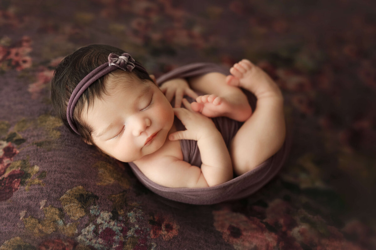 Newborn baby girl  laying on her back curled up in a purple swaddle on a purple floral backdrop at her newborn session with a photographer at a great falls va photo studio