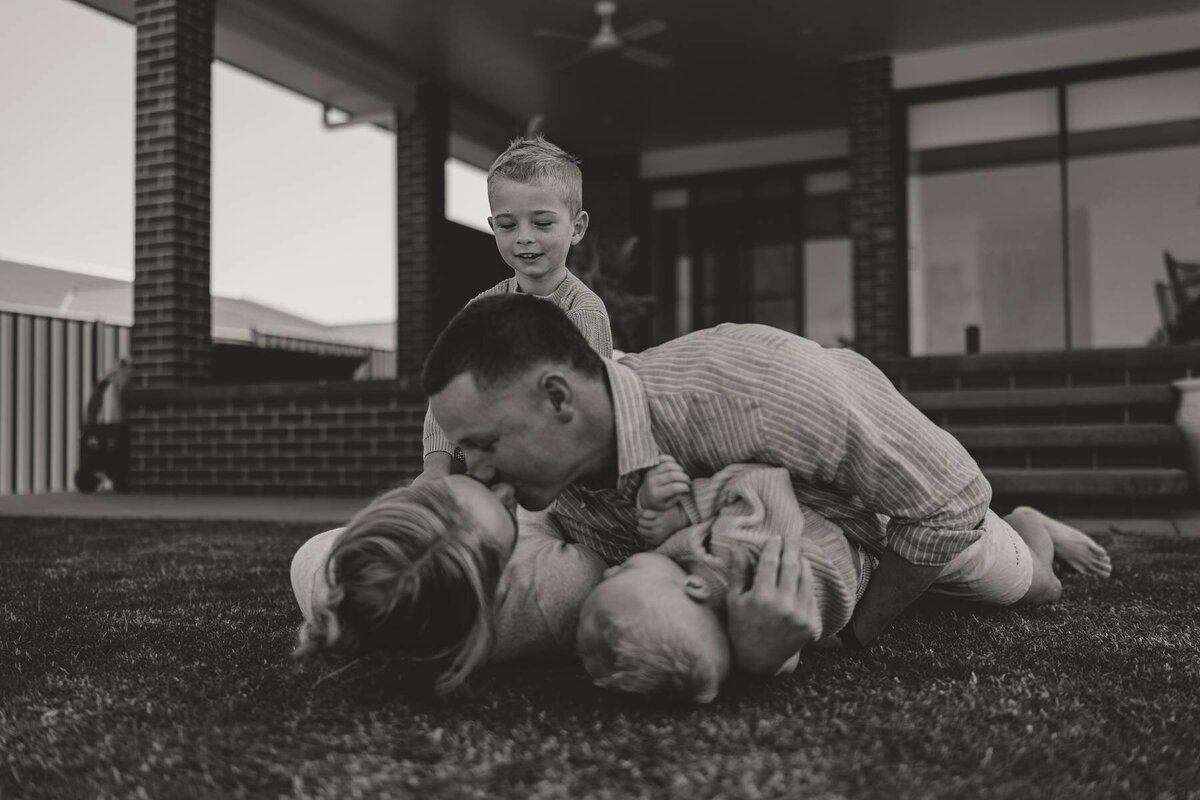 Candid family photography capturing a father playing on the lawn with his three young children.