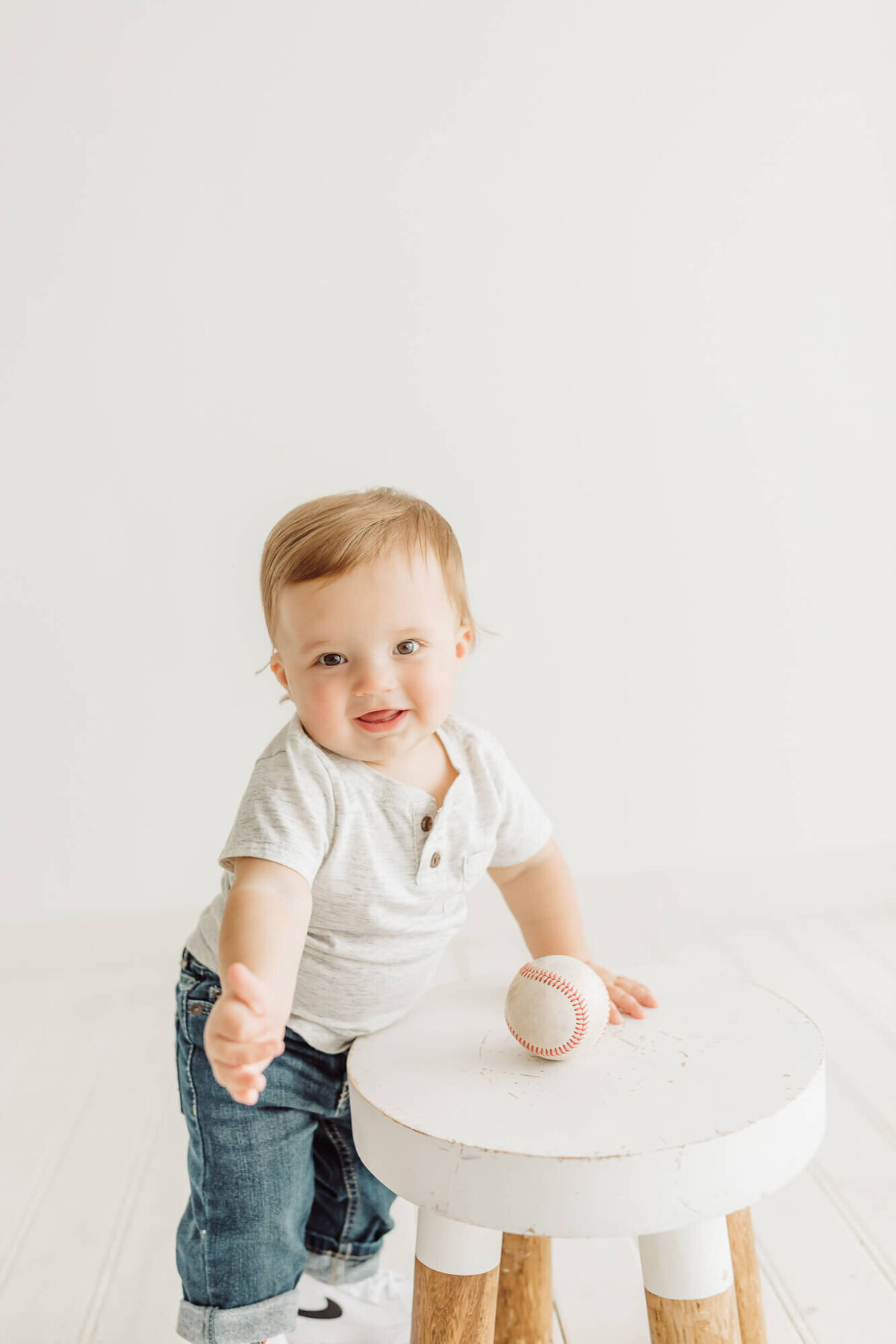 One year old little boy standing against a bench with baseball for his studio session by Ally's Photography.