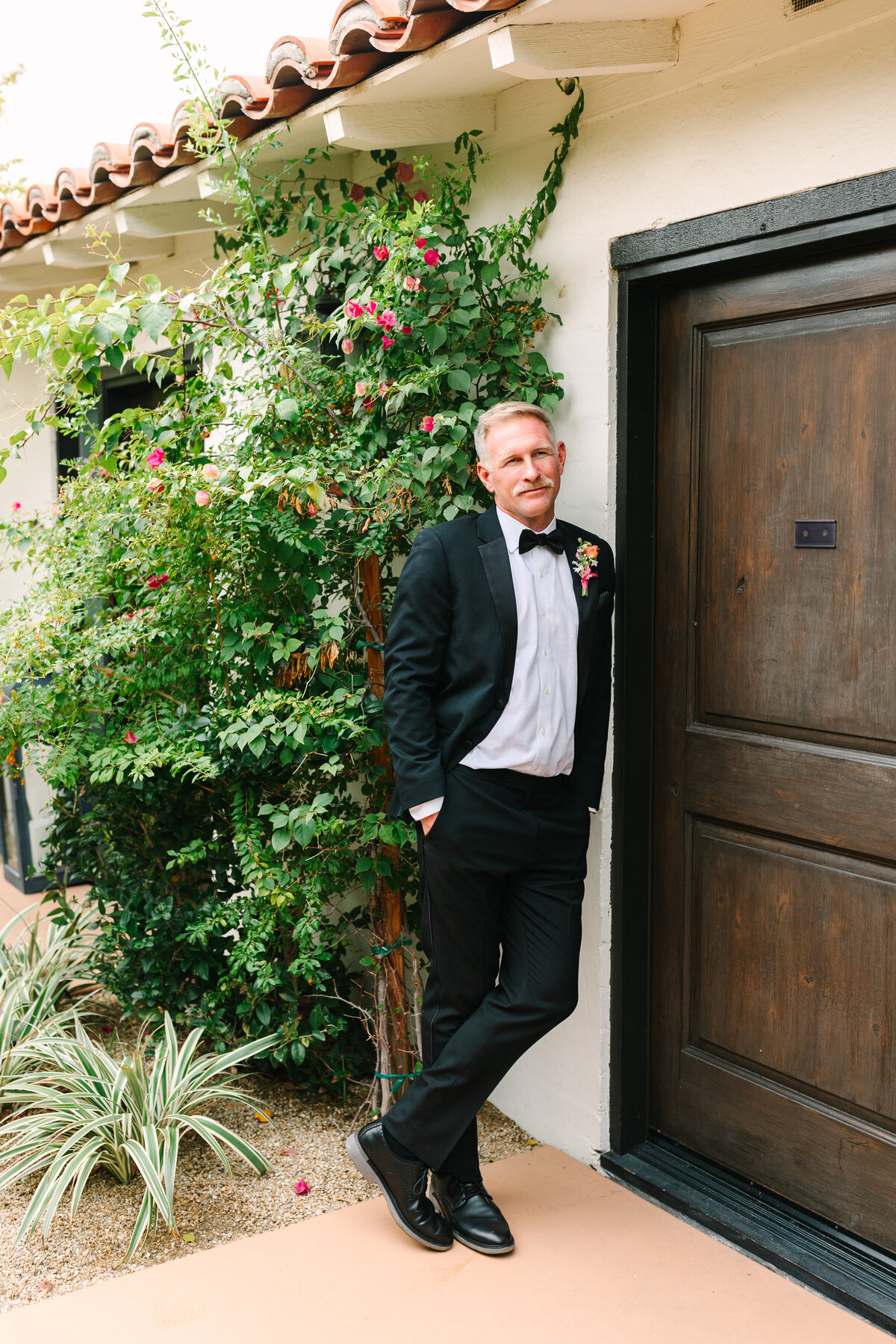 groom casually leaning against side of a building in napa with greenery beside him and dressed in tuxedo.