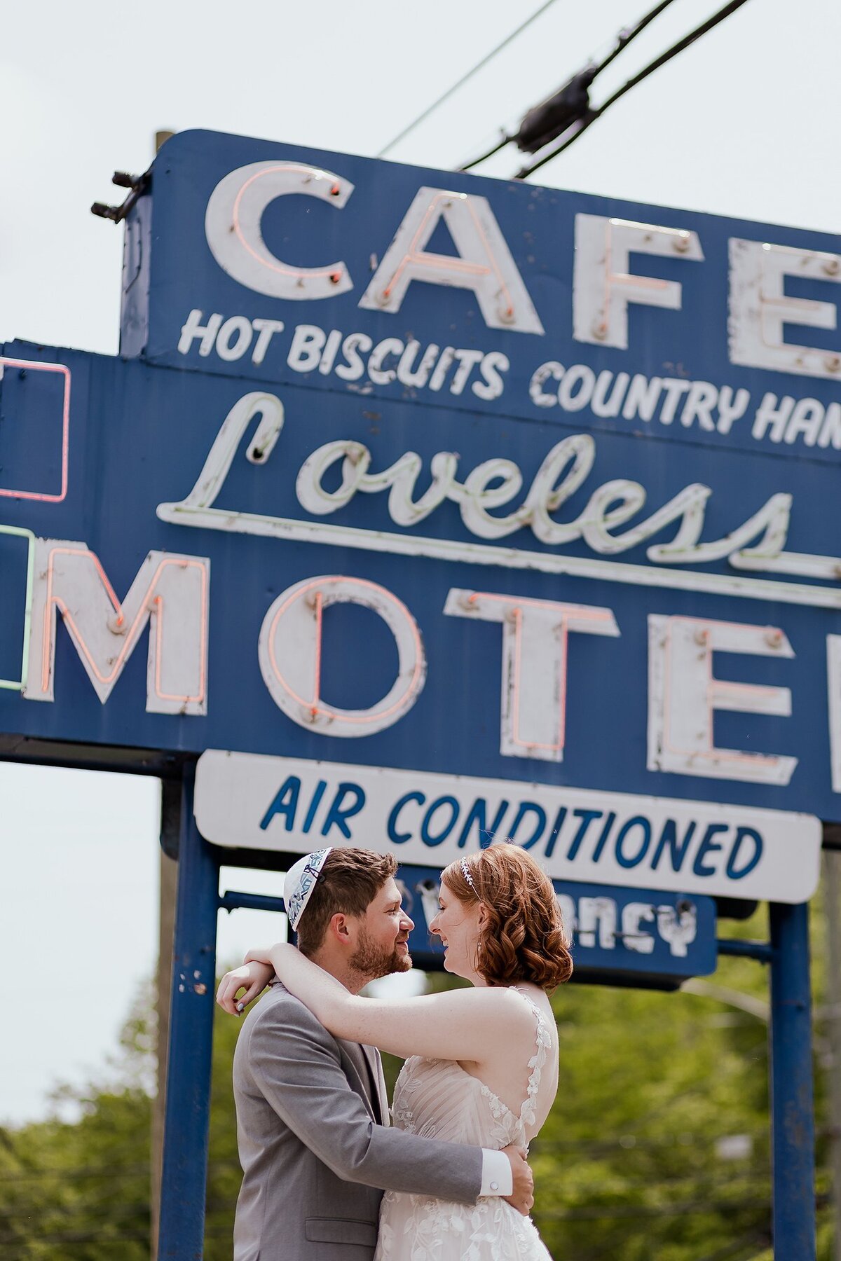 Jewish bride wearing a white v-neck gown with a low back embraces the groom wearing a white and blue yarmulke and a light gray suit in front of the Loveless Motel sign at Loveless Cafe in Nashville at their Jewish wedding