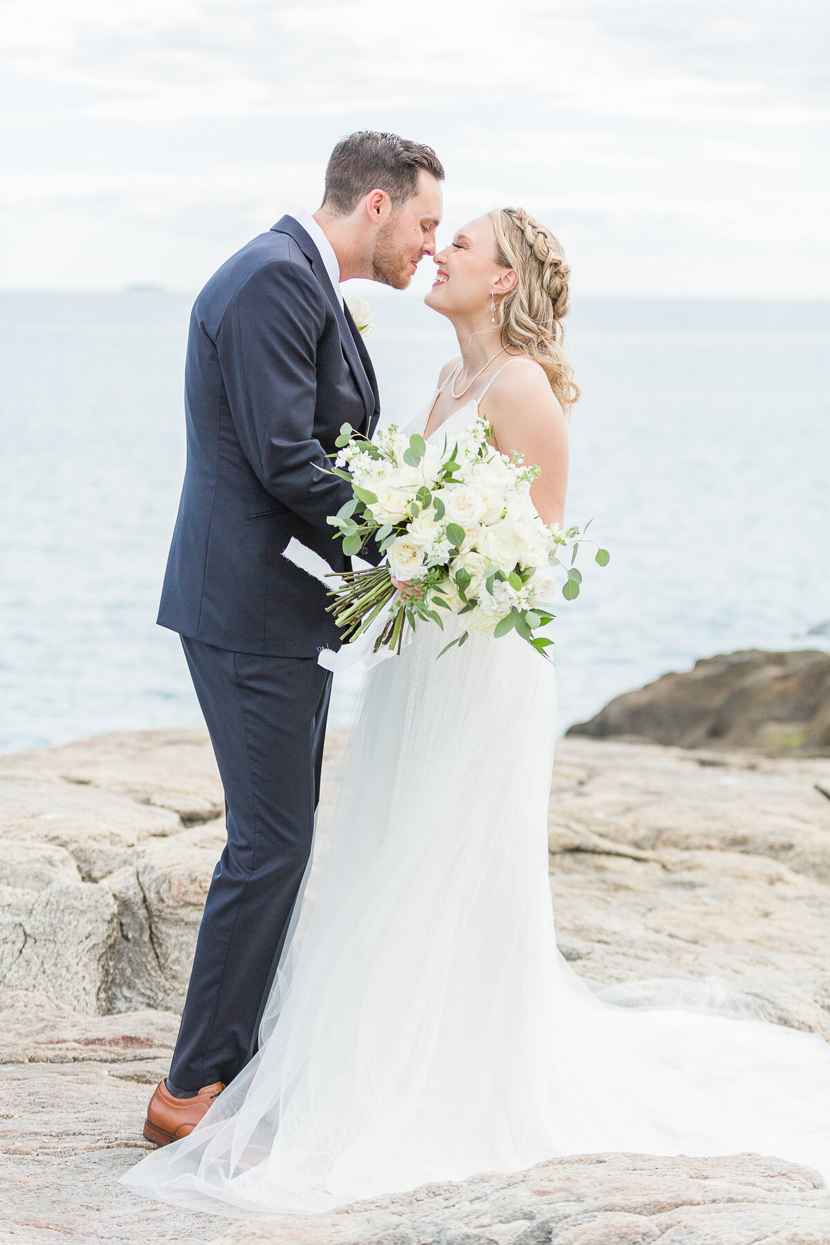 Bride and groom stand facing each other on the rocks on the shoreline for their Madison Beach Hotel Wedding in Madison, CT. The are smiling at each other and touching noses. Captured by best New England wedding photographer Lia Rose Weddings.