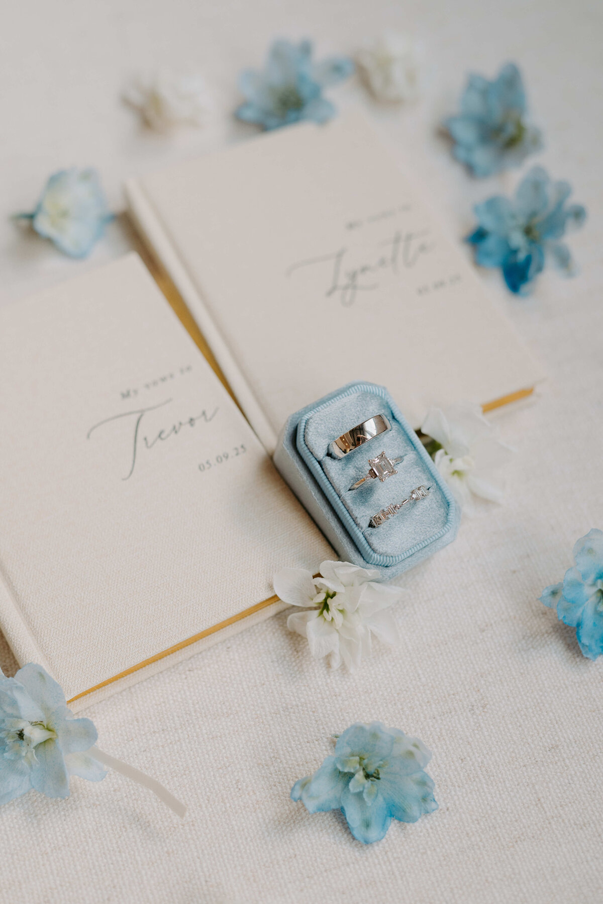 Elegant wedding scene with two vow books on a textured surface, flanked by blue flowers. A light blue box displays three silver rings, evoking romance and commitment.