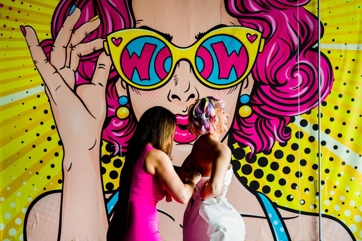 A woman helping a bride zip her dress in front of a bright mural.