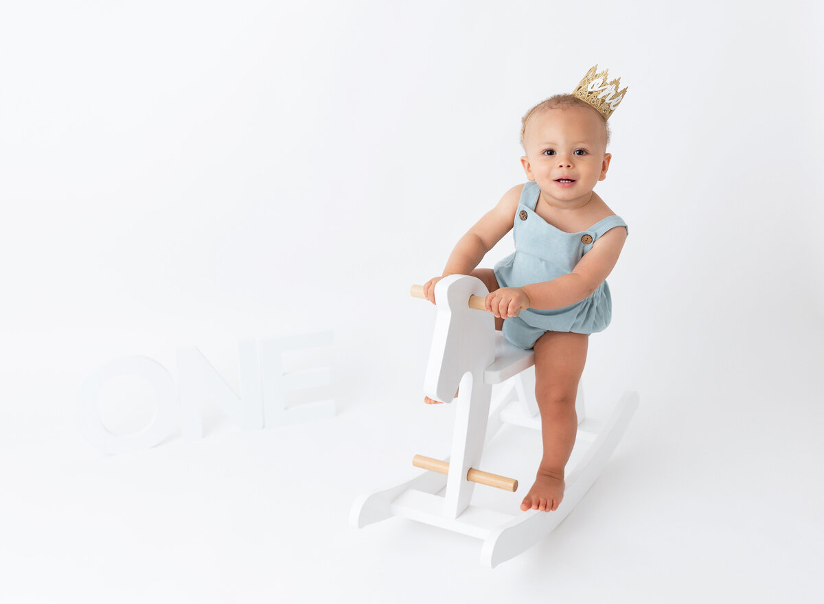 Baby boy wearing light blue outfit and sitting on white horse at first birthday photo session