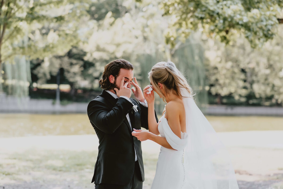 A bride and groom in wedding attire stand tearfully facing each other in a sunlit park. The scene is emotional and intimate, with a soft, natural background.