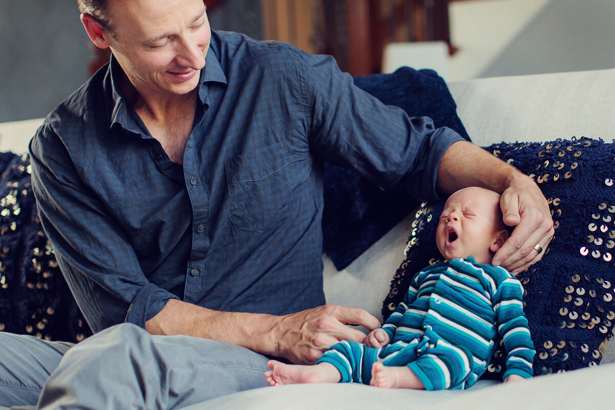Newborn boy in blue stripes is sitting up with Dad's assistance, big yawn, as Dad smiles at him.