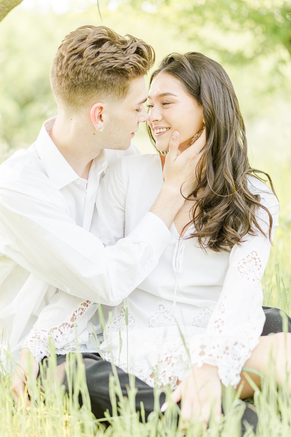 Couple are sitting in the grasses at Smolak Farm for their engagement photoshoot. One partner is touching the other's cheek and bringing them in for a kiss. Captured by Lia Rose Weddings