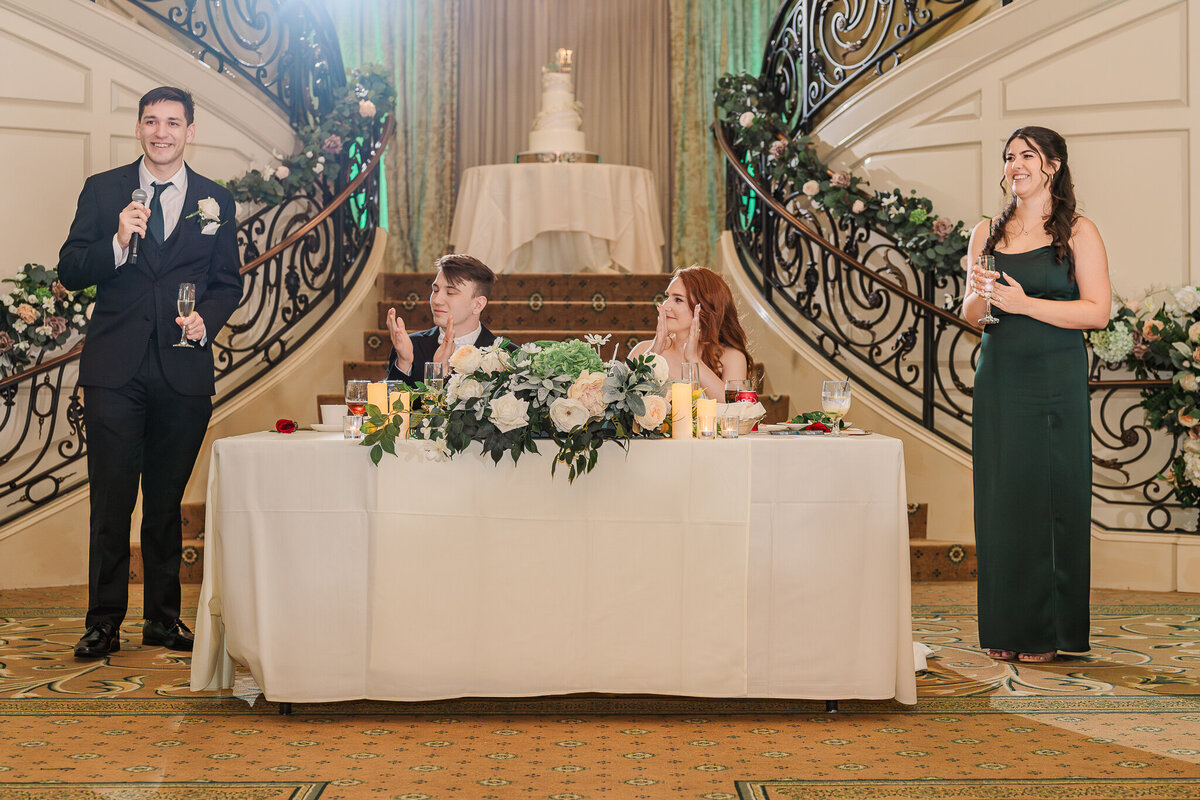 The best man and maid of honor giving speeches and toasts to the bride and groom under the Grand Staircase at The Prestonwood Country Club while enjoying their Raleigh wedding photography session by JoLynn Photography