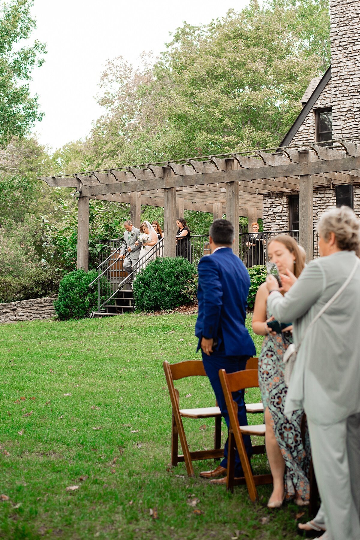 The bride wearing a sleeveless lace wedding dress is escorted to the wedding ceremony by her father wearing a light gray suit at the Stone House at Arrington Vineyard. The wedding guest stand from the garden wood chairs  and look towards her.