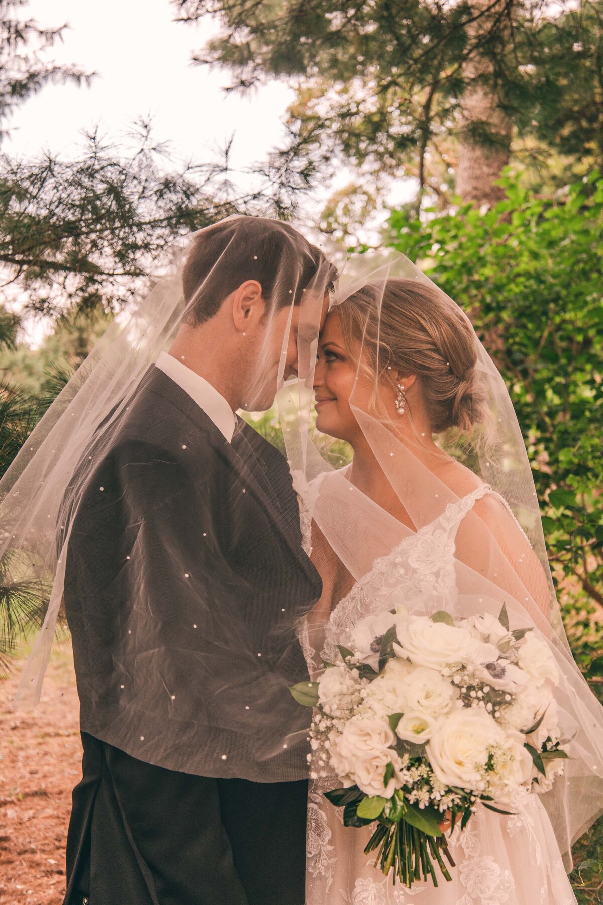 A bride and groom standing close under a veil in a wooded area at Park Farm Winery, the bride holding a bouquet of white flowers, both smiling gently at each other.