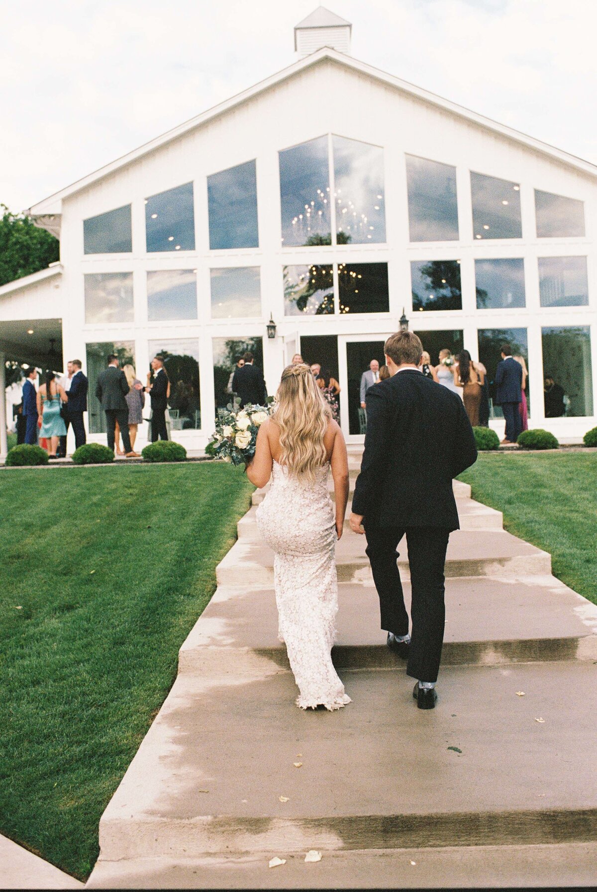 A couple in formal attire walks hand-in-hand up a path to a modern, white building with large windows. Guests in elegant clothing talk outside, celebrating.
