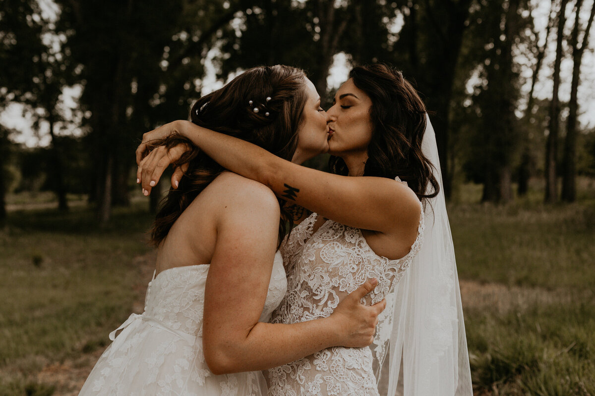 two brides kissing in front of tall green trees