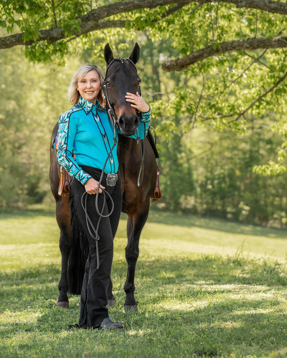 blonde female equestrian wearing a blue western show shirt and black chaps, standing under a gorgeous tree with her dark bay quarter horse mare. they are surrounded by green trees and pasture.