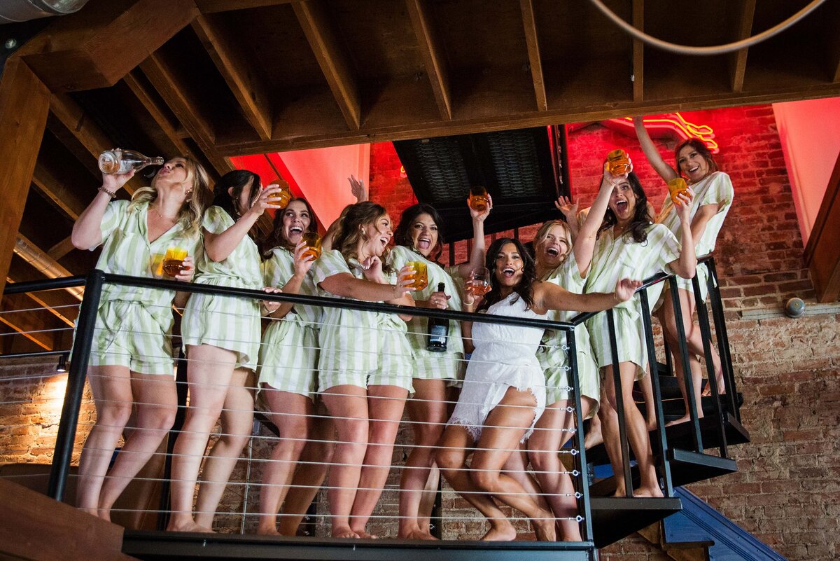 A bride and her bridesmaids dressed in pajamas stand on a balcony and celebrate while sipping glasses of champagne.