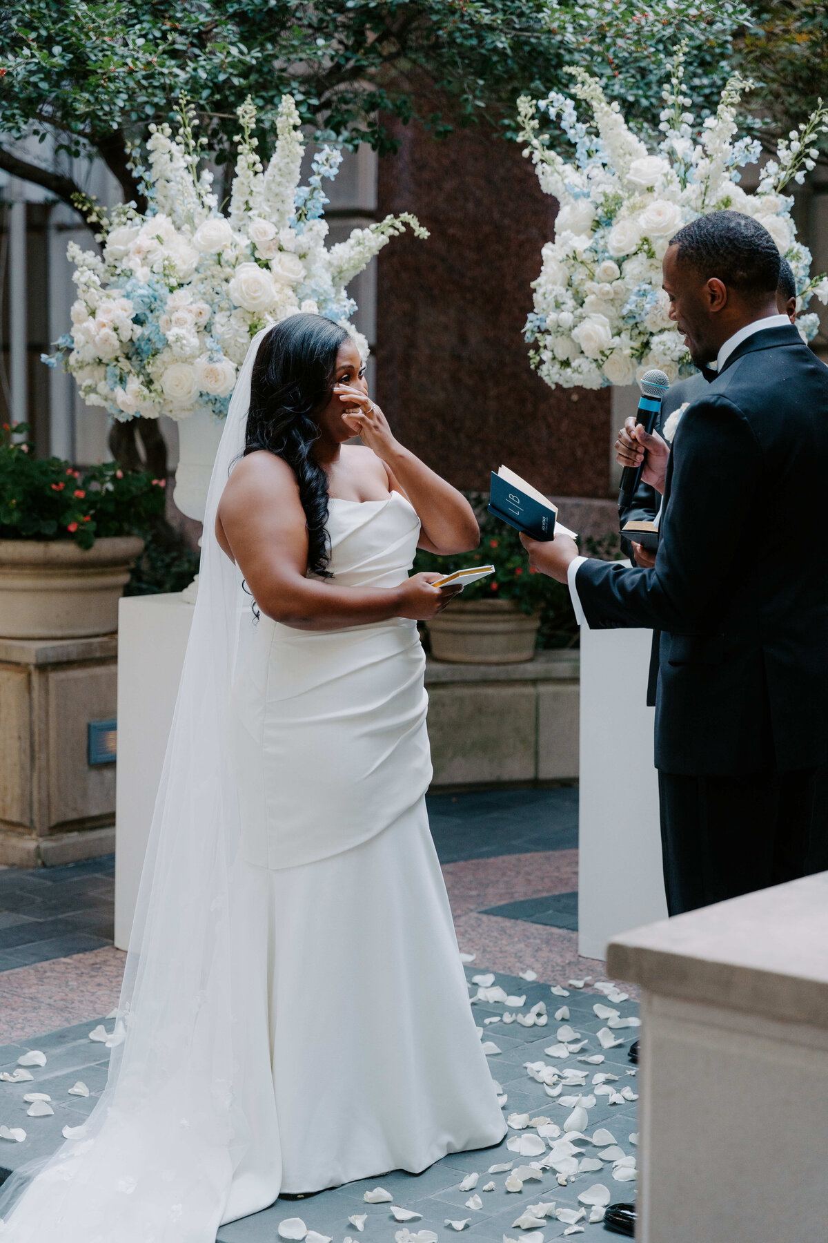 A bride in a white dress smiles emotionally, facing a groom in a black suit holding a microphone and book. They're surrounded by white flowers outdoors.
