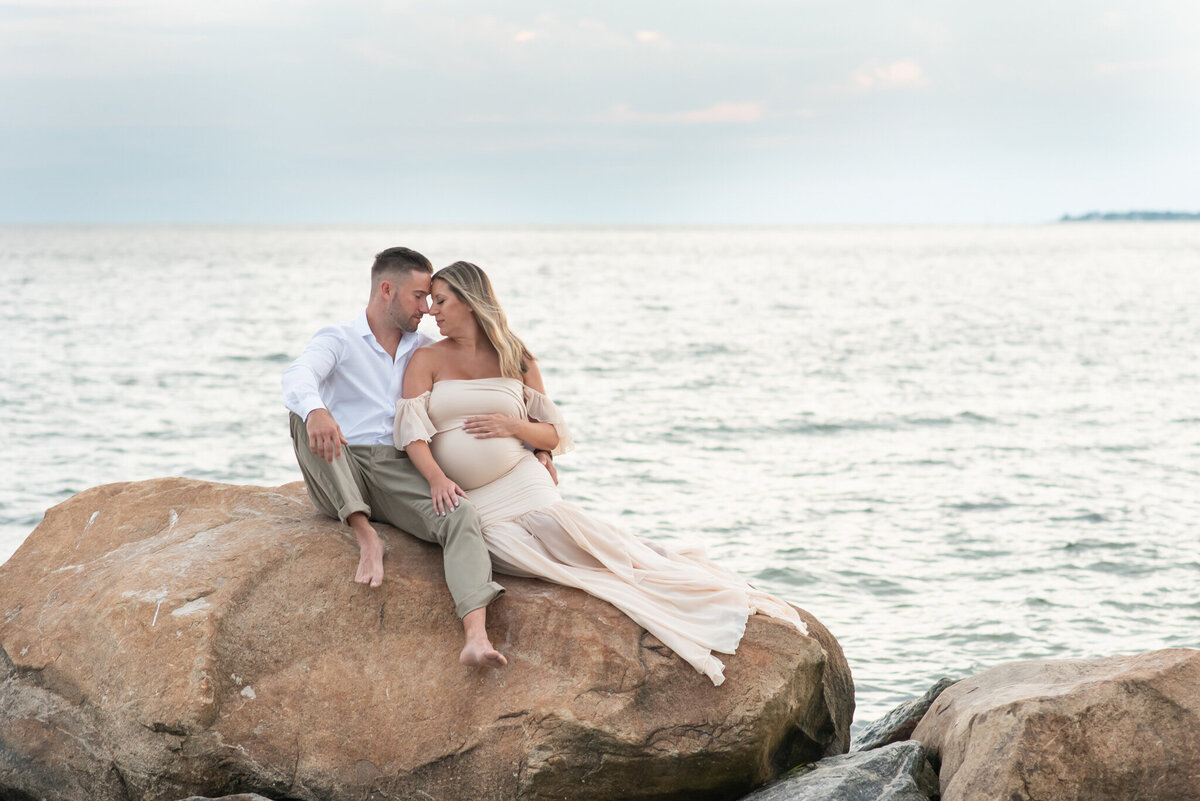 Expecting couple sitting on a rock on the beach at sunset in Waterford, Connecticut