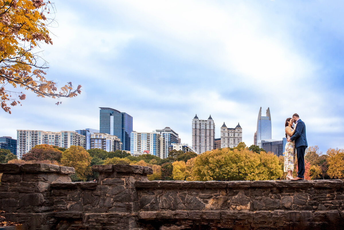 Engaged-couple-standing-on-a-rock-wall-with-their-foreheads-together-with-the-downtown-Atlanta-city-skyline-in-the-background-at Piedmont-Park