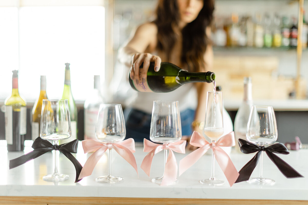 Photo of Sip and Savors owner pouring wine into glasses dressed with satin ribbons for a brand session.