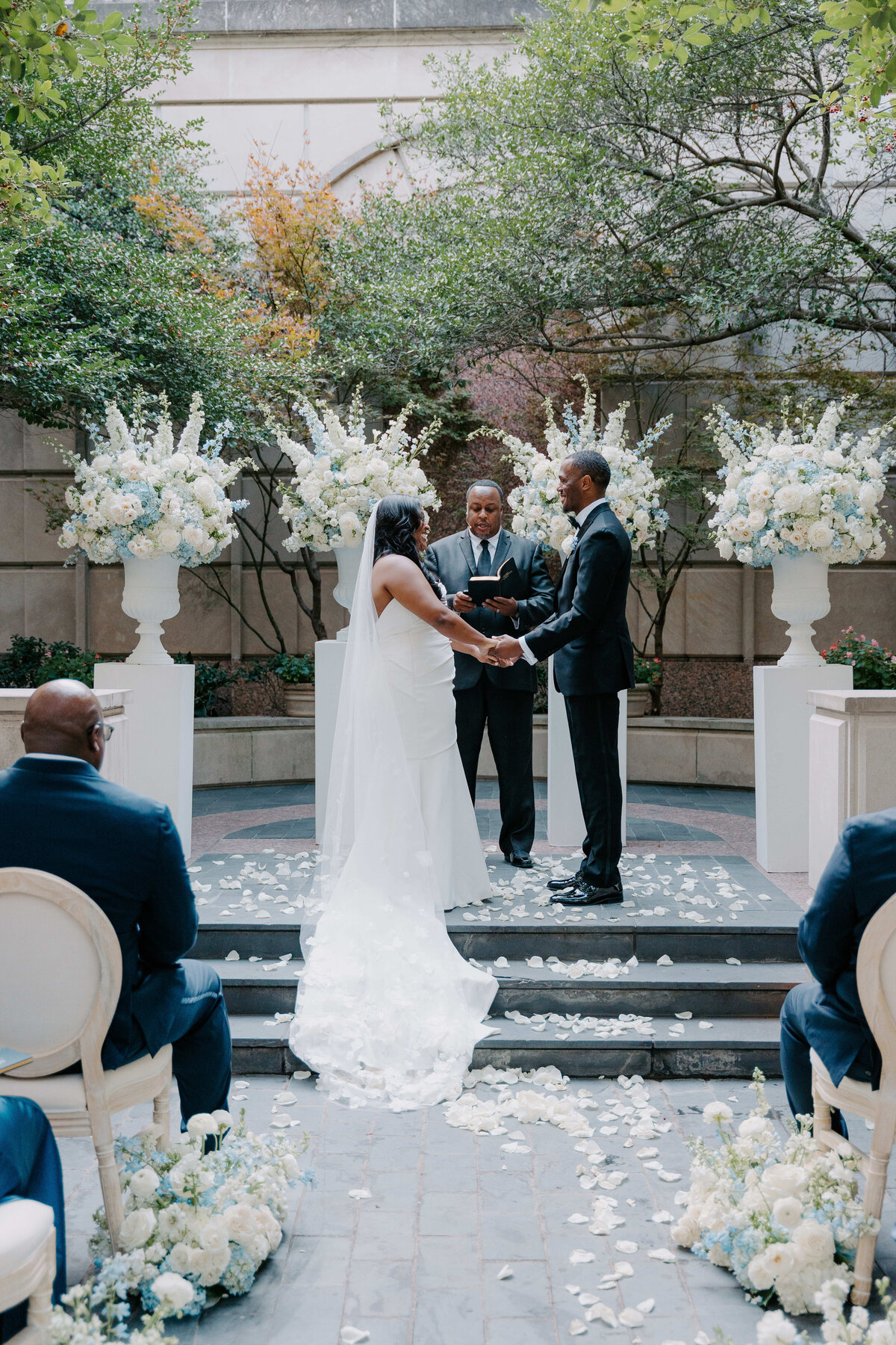 Bride and groom exchange rings at a wedding ceremony, with an officiant standing between them. They are surrounded by white floral arrangements, exuding joy.