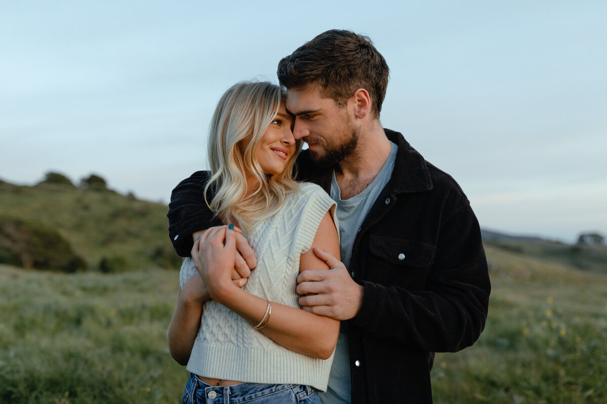 Man hugging woman from behind while they look into each other's eyes and smile with green hills in background