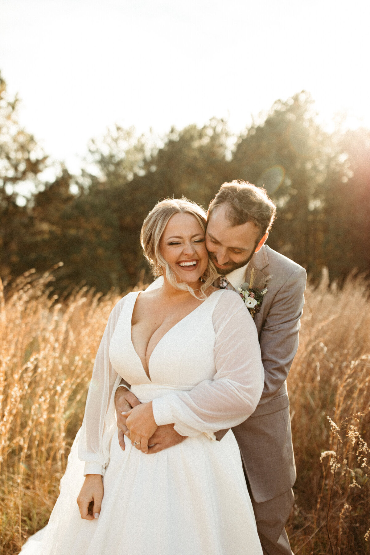 Groom squeezing bride from behind and making her laugh in a field with tall grass