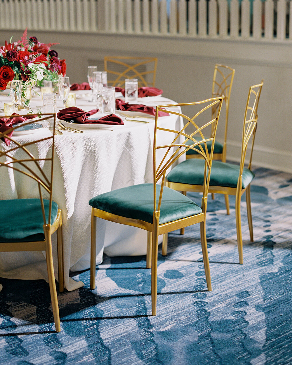 Luxurious table decor for a December wedding at the Don Cesar Hotel in St. Pete Beach, FL. Features green velvet chairs with gold backs, white linen tablecloths, red napkins, and a red floral centerpiece in a gold vase.