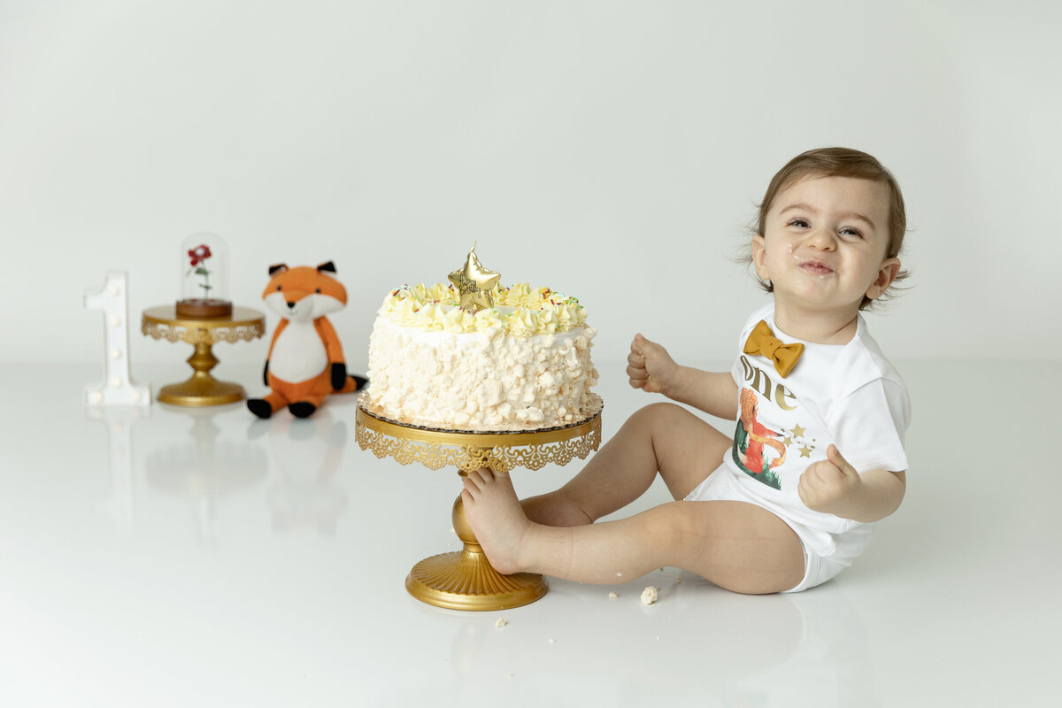 A toddler boy in a "one" onesie happily eats some cake in a photo studio