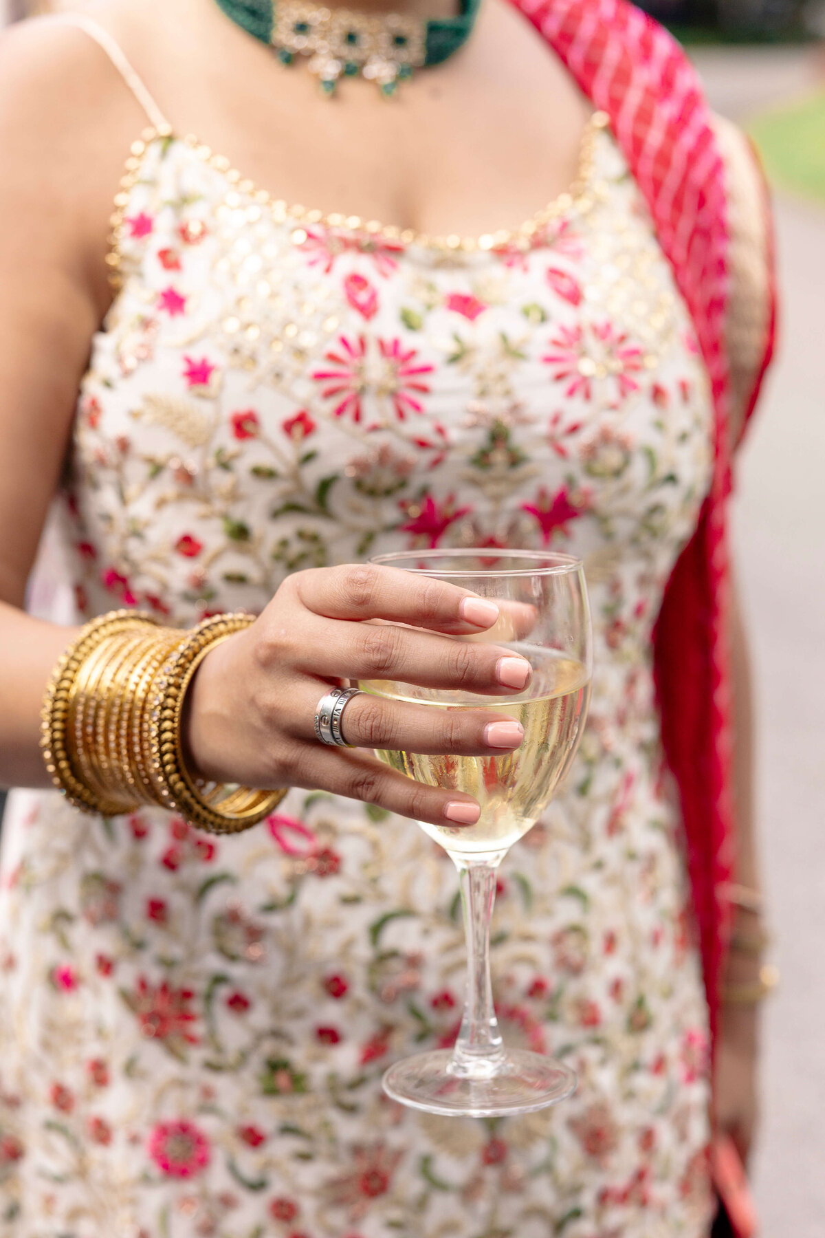 A woman in a floral dress holds a glass of white wine. She wears gold bangles and rings, and a red dupatta draped over her shoulder. Her outfit is adorned with intricate embroidery, and shes accessorized with a green choker necklace.