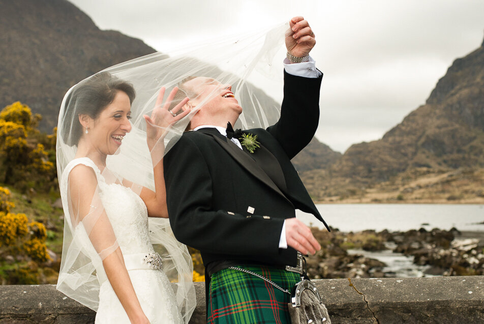 brunette bride wearing an a-line wedding dress with capped sleeves with her husband wearing a scottish kilt wedding suit, pulling the bride's veil off his head while standing on a stone bridge at the Gap of Dunloe