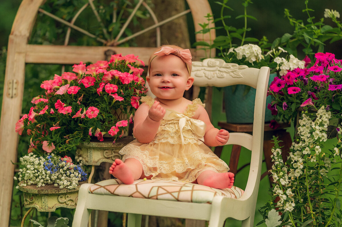A baby girl sits in an outdoor, vintage-inspired portrait set surrounded by pink flowers.