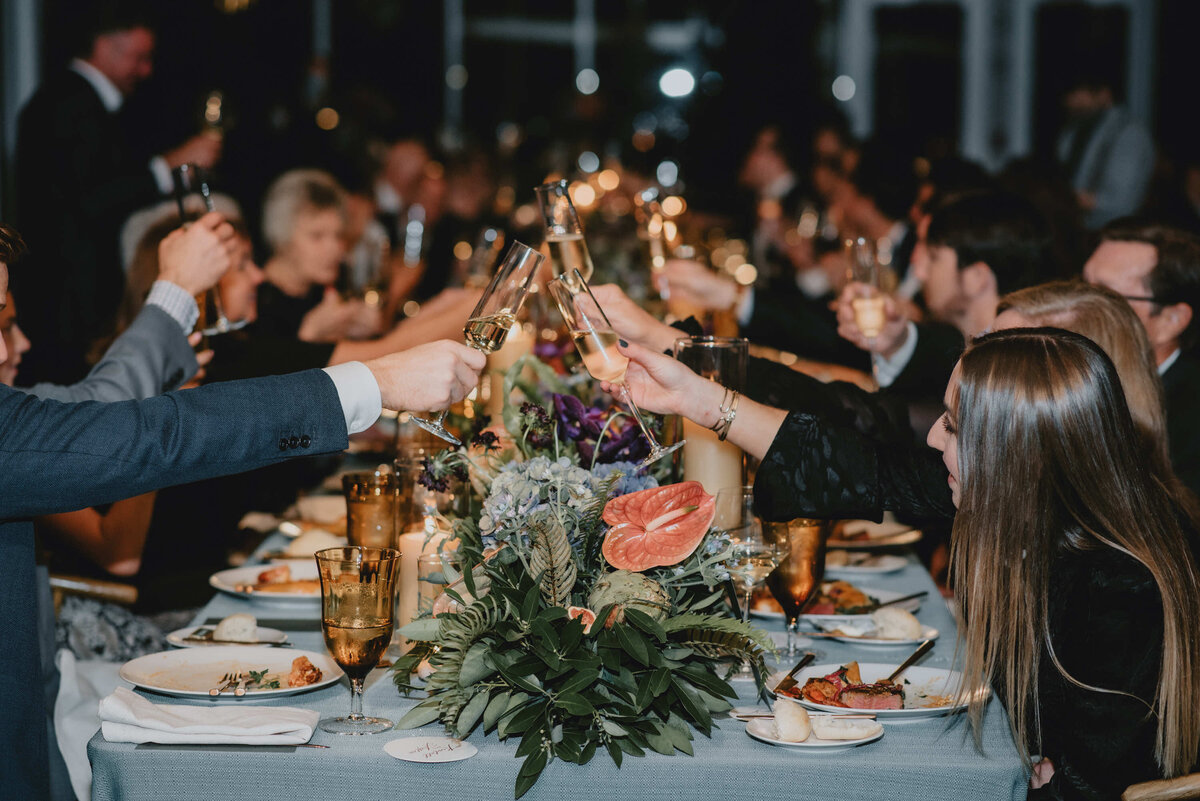 A large group gathers around an elegant, candlelit dinner table, raising glasses for a toast. The mood is celebratory, with floral centerpieces and warm lighting.