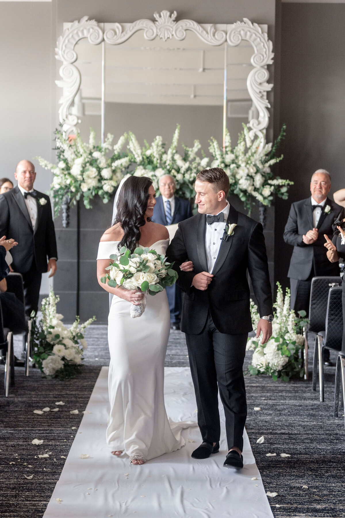 A bride in an off-shoulder gown and a groom in a tuxedo walk down the aisle, smiling. They are surrounded by guests in formal attire, with a white floral arrangement and a large ornate mirror in the background.