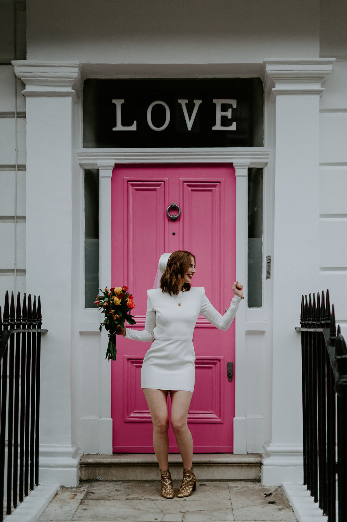 A bride wearing a Neta Porter dress dances outside a pink door in Chelsea, London. She is wearing her mums vintage hat, has red lipstick and a bouquet she picked up that morning from Chelsea Flower Show.