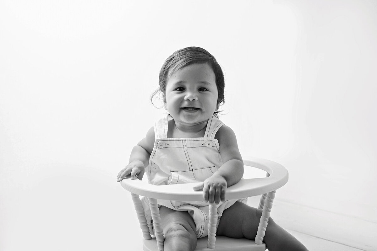 A young toddler smiles for a Lafayette Baby Milestone Photographer while sitting in a baby chair in overalls in black and white