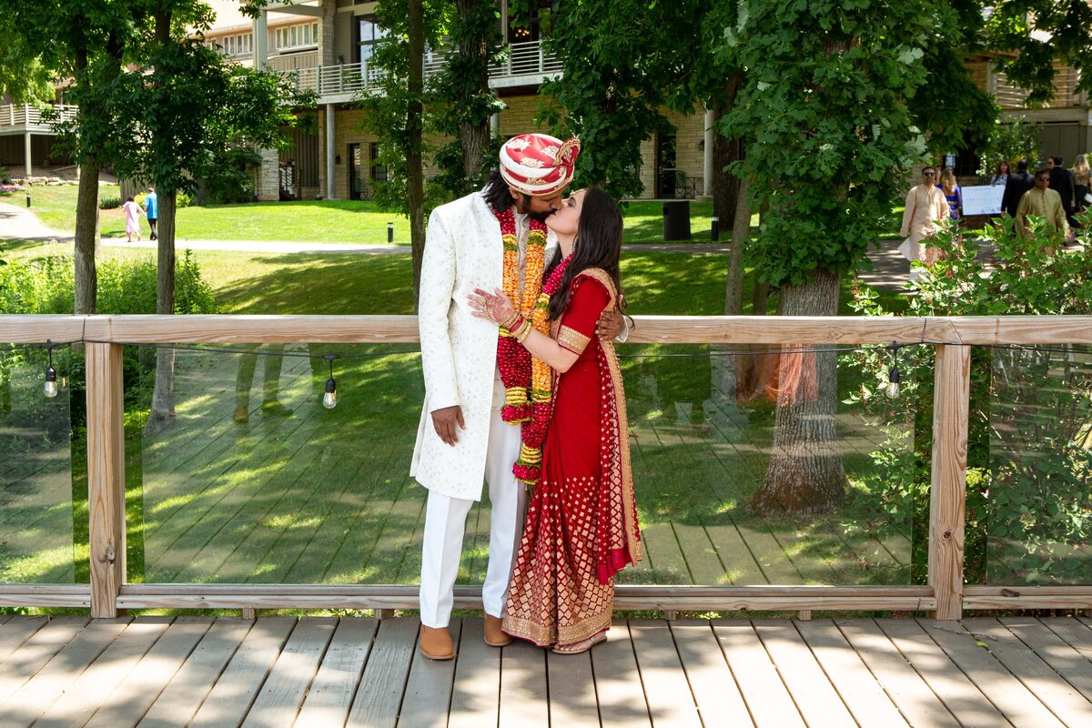 A couple in traditional Indian wedding attire, coordinated by a top Des Moines wedding planner, sharing a kiss on a wooden bridge in a park setting.