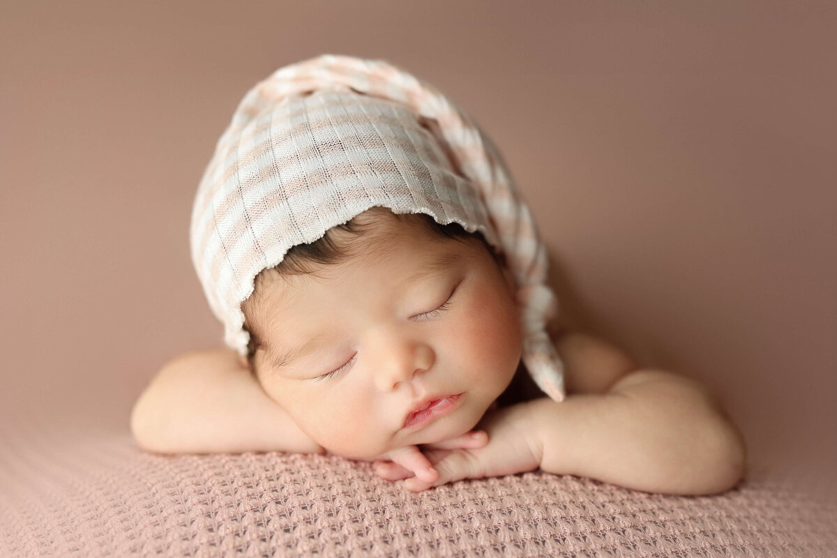 baby girl laying on her hands with a striped sleepy cap on a pink backdrop in a newborn session