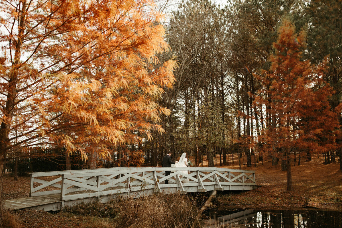 Wide angle shot of a bride and groom walking across a bridge over a pond with orange fall colored leaves around them