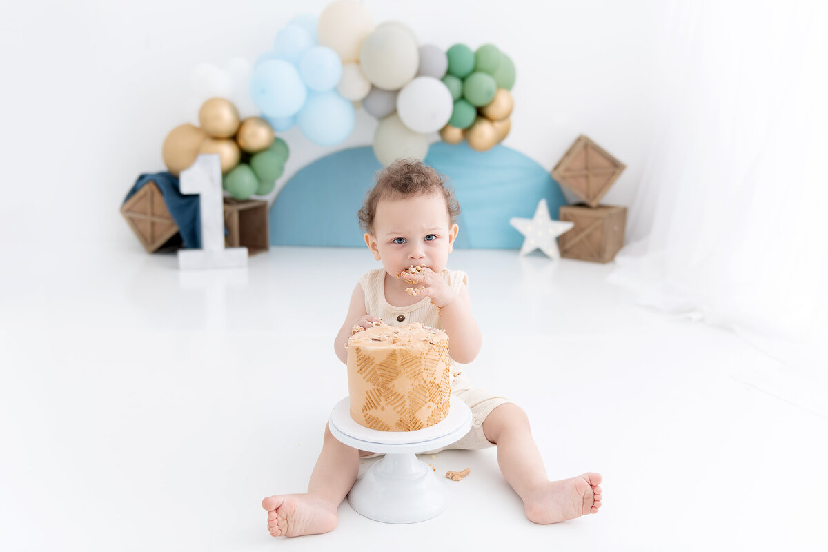 A toddler boy in a cream onesie sits on the floor of a studio eating a cake decorated by a childrens photographer Atlanta