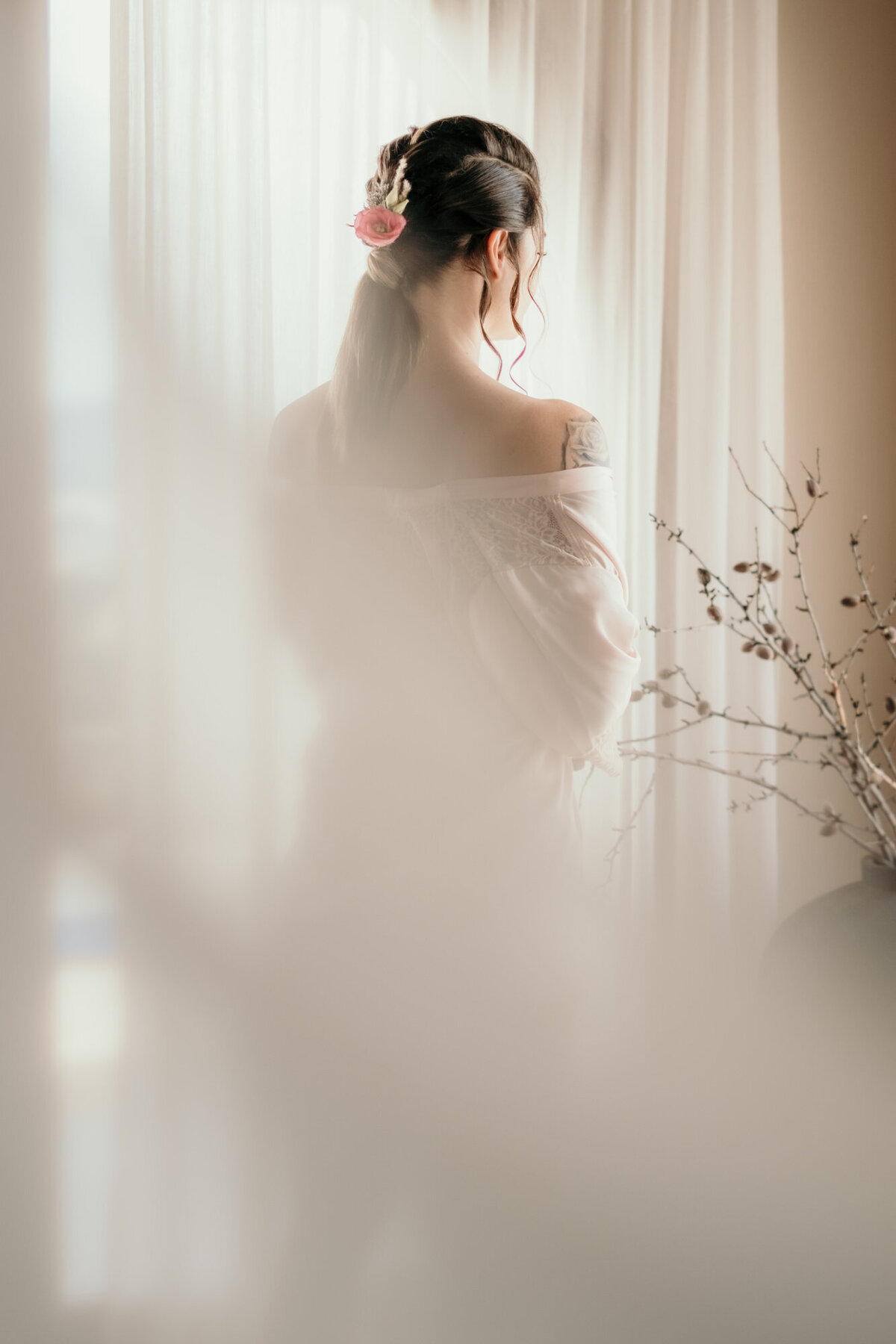 In einem Morgenmantel steht die Braut mit Blumen im Zopf mit dem Rücken zur Kamera.