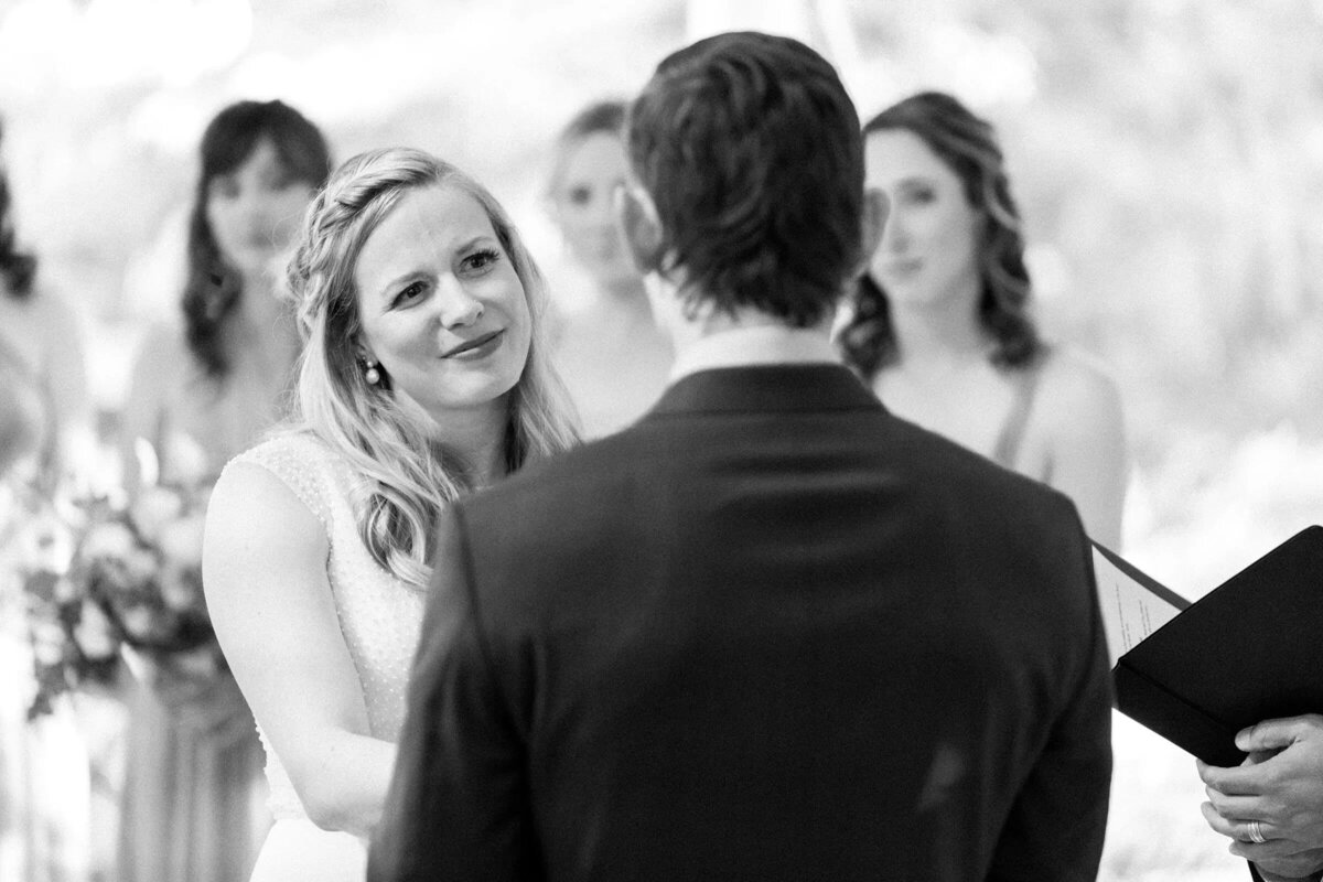 A black and white photo capturing a bride looking lovingly at her groom during their wedding ceremony with a soft focus on the guests in the background.