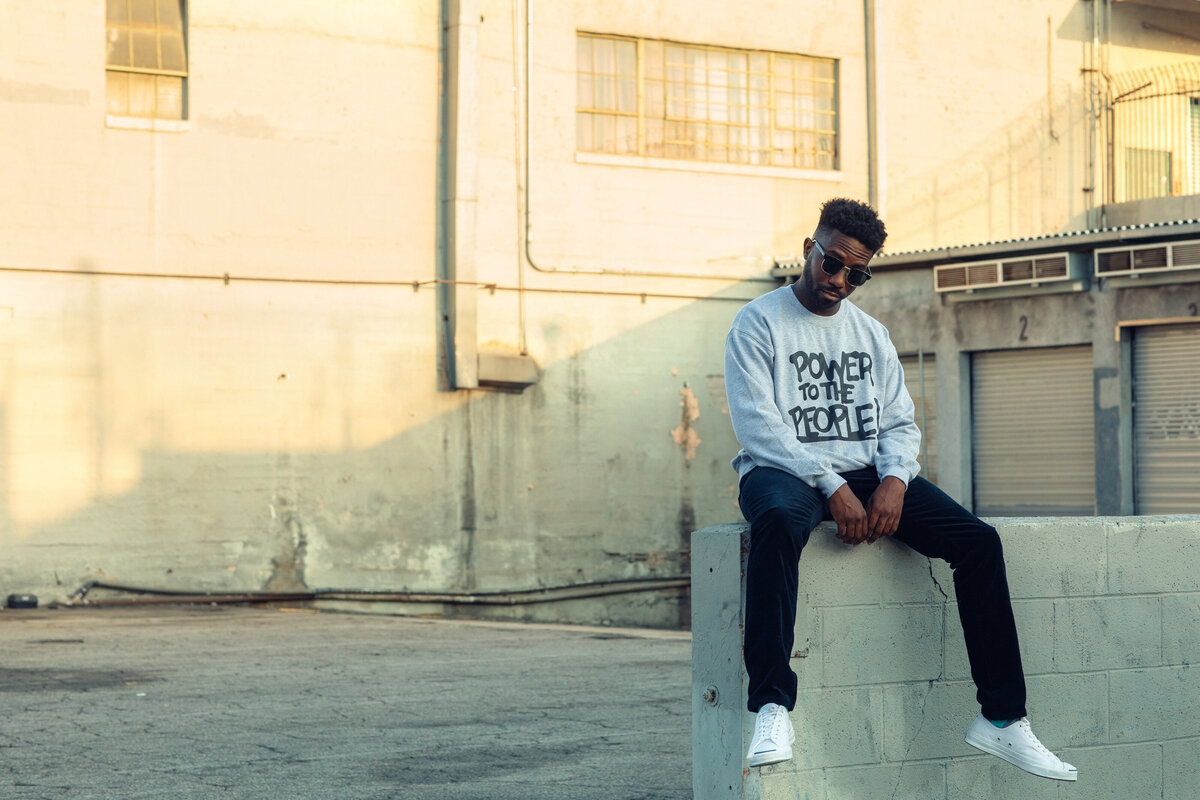 Portrait Photo Of Young Black Man In Gray Sweater Sitting On Top Of a Wall Los Angeles