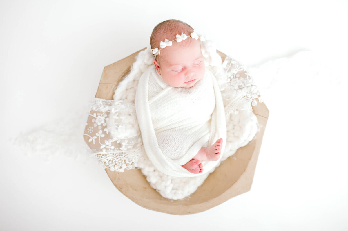 Baby wrapped in white swaddle, sleeping in a bowl with lace drapped over her
