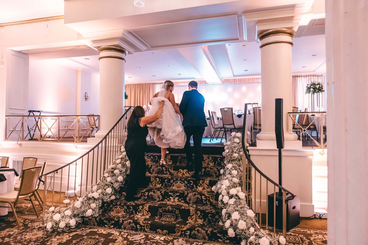Bride and groom climbing a staircase decorated with white flowers, helped by two people, in an elegantly lit reception hall at an Iowa wedding.