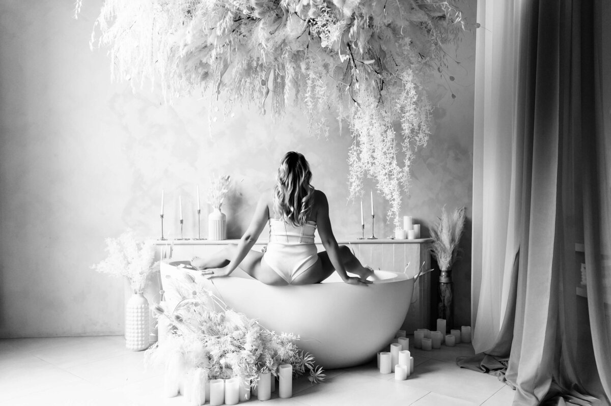 Black and white photo of a woman sitting in a bathtub her legs on the side with pampas all around at Mint Room Studios Toronto.
