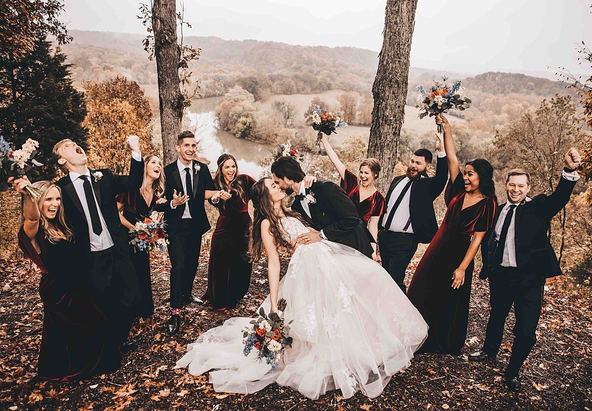 Bride in a long white gown and veil being dipped and kissed by the groom in a black suit on a Tennessee mountain top while the bridal party in maroon and black cheer them on