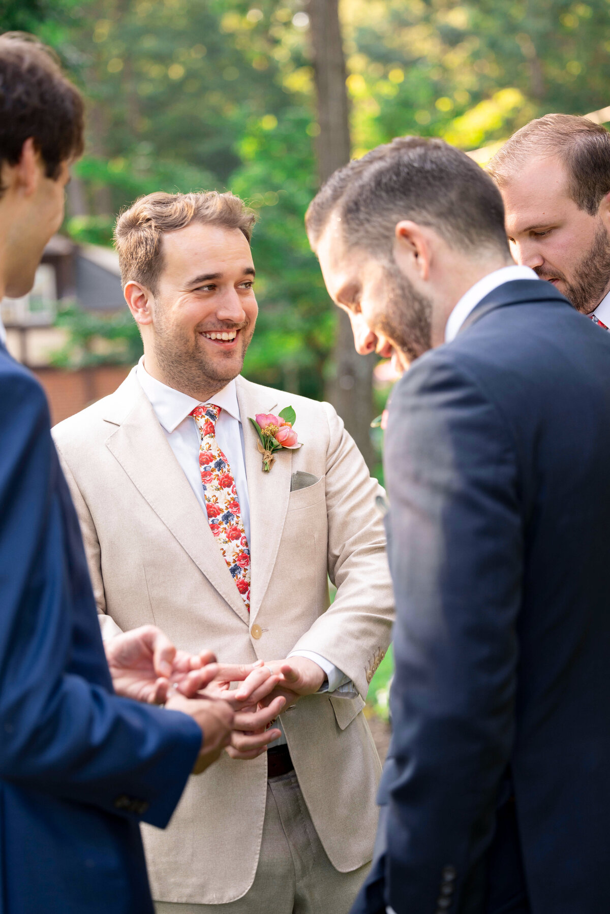 A group of men in suits stand outdoors in a lush green setting, likely at a wedding. One man in a cream suit and floral tie smiles and holds out his hand showing off his wedding ring, surrounded by others in dark suits, all engaged in conversation.